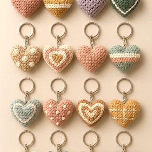 Puede incluir: Una colección de dieciséis llaveros de corazón de ganchillo en varios colores, incluyendo rosa, amarillo, morado y verde. Cada corazón está unido a un llavero plateado. Los corazones presentan diferentes patrones y diseños.
