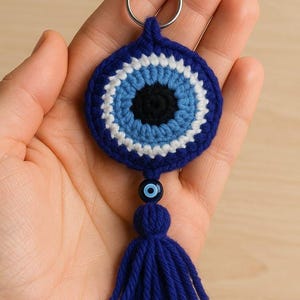 Könnte beinhalten: Ein handgefertigter blauer Häkel-Schlüsselanhänger mit einem bösen Blick-Design. Der kreisförmige Anhänger hat eine schwarze Mitte, umgeben von hellblauen, weißen und dunkelblauen Ringen. Ein passender blauer Quastenanhänger und ein silberner Schlüsselring vervollständigen das Design.
