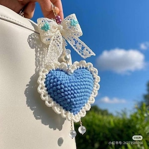 Op de afbeelding: Een handgemaakte sleutelhanger met een blauw gehaakt hart en een witte geschulpte rand. De sleutelhanger heeft een witte kanten strik met turquoise bloemaccenten en bungelende heldere kralen. De achtergrond toont een helderblauwe lucht.