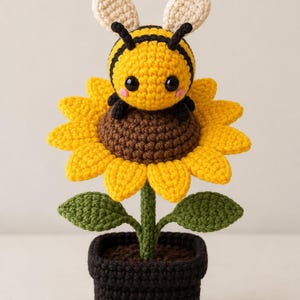 Puede incluir: Un girasol de ganchillo con una abeja sonriente encima. El girasol tiene pétalos amarillos, un centro marrón, hojas verdes y un tallo verde. La abeja es amarilla y negra con antenas blancas. La flor está en una maceta negra.