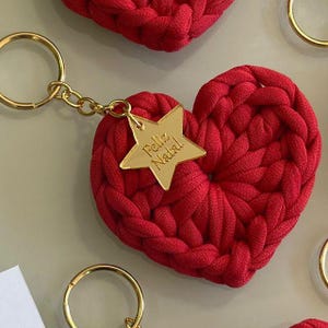 May include: A red, heart-shaped keychain made of a thick, woven material. A gold-colored star charm with the words "Feliz Natal" is attached. The keychain has a gold-colored ring. The background is a light, neutral color.