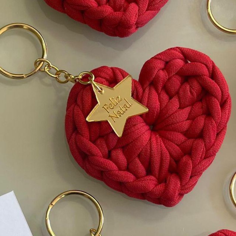 May include: A red, heart-shaped keychain made of a thick, woven material. A gold-colored star charm with the words "Feliz Natal" is attached. The keychain has a gold-colored ring. The background is a light, neutral color.