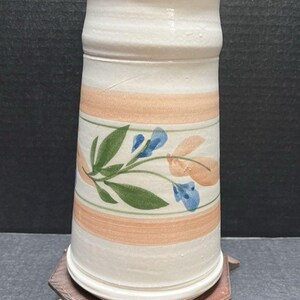 Könnte beinhalten: Eine zylindrische Keramikvase in cremeweißer Grundfarbe. Die Außenseite ist mit horizontalen pfirsichfarbenen Bändern und einem zentralen, handgemalten Blumenmotiv verziert, das aus grünen Blättern sowie blauen und pfirsichfarbenen Blüten besteht.