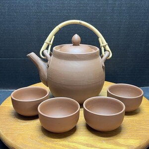 以下が含まれることがあります： 竹製の持ち手と蓋が付いた丸い急須と、持ち手のない小さな湯呑み4つで構成されたテラコッタ製の茶器セット。全体的にマットなアースブラウンの仕上げが施されており、シンプルで機能的なデザインが特徴です。