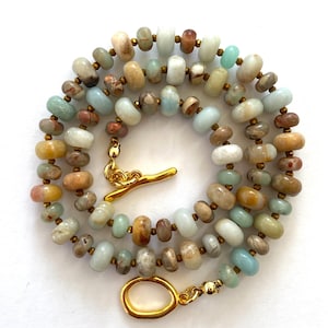 Puede incluir: Collar de varias hebras con cuentas ovaladas multicolores en tonos beige, marrón y azul verdoso. El collar se cierra con un cierre dorado. Las cuentas son de diferentes tamaños.