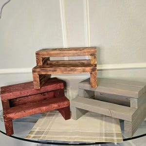 Könnte beinhalten: Drei Holz-Trittschemel in verschiedenen Farben: ein dunkles Rotbraun, ein helles Grau und ein mittleres Braun mit verbranntem Finish. Jeder Hocker hat zwei Stufen und ein einfaches, robustes Design. Sie werden auf einem Glastisch ausgestellt.