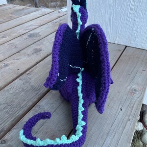 May include: A handmade crochet dragon in shades of purple, navy blue, and light blue. The dragon has large wings, a long tail, and two upright horns. The dragon is sitting on a wooden surface.