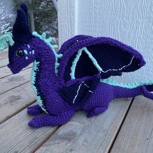 May include: A handmade crochet dragon in shades of purple, navy blue, and mint green. The dragon has large wings, a long tail, and detailed features. The dragon is lying on a wooden surface.
