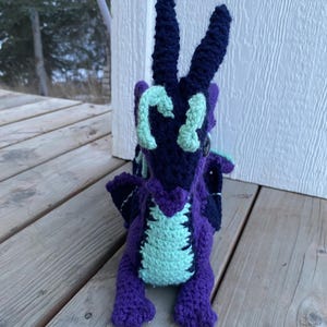 May include: A handmade crochet dragon in shades of purple, navy, and teal. The dragon has large horns, wings, and a detailed body. The dragon is sitting on a wooden surface.