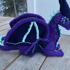 May include: A handmade crochet dragon in shades of purple and navy blue, with light blue accents on the wings and spine. The dragon has large, dark eyes and is posed on a wooden surface. A whimsical, fantasy-themed item.