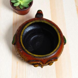 Pode incluir: Um vaso de cerâmica redondo, feito à mão, com interior escuro e exterior texturizado em tons de vermelho, laranja e amarelo. Uma pequena planta suculenta num vaso castanho está por perto. O vaso tem um design decorativo único.