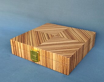 Op de afbeelding: Vierkante houten doos met een geometrisch patroon op het deksel. De doos is gemaakt van lichtbruin hout met donkerbruine strepen. Hij heeft een goudkleurige sluiting. De doos staat op een blauwe achtergrond.