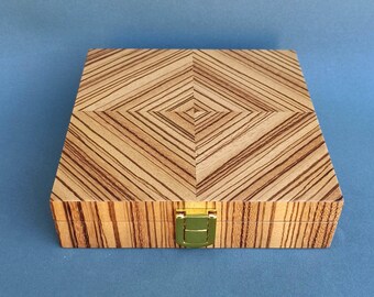 Op de afbeelding: Een houten kist met een geometrisch patroon op het deksel. De kist is gemaakt van lichtgekleurd hout met donkerbruine strepen. Hij heeft een goudkleurige sluiting en staat tegen een blauwe achtergrond.