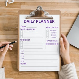 May include: A clipboard with a "DAILY PLANNER" sheet, featuring sections for date, priorities, to-do list, meal tracker, and water intake. A pencil rests on the wooden desk. A laptop and earbuds are also visible.