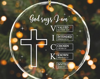 May include: Clear glass Christmas ornament with the text "God says I am" and a white cross. The ornament lists the words "Valued, Intended, Chosen, Known, Invited" with corresponding Bible verses. A gold string is attached for hanging.