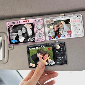 May include: Three camera-shaped car visor clips, each displaying a photo. One is pink with the text "Favorite Person." Another is white with "ILOV3U" and "Drive Safe." The third is black with "Get Home Safe."