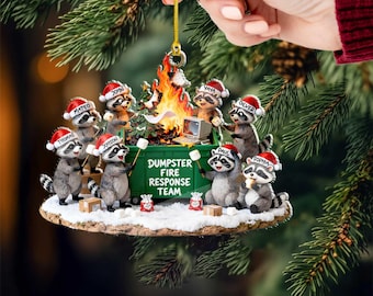 May include: A Christmas ornament featuring a group of raccoons wearing Santa hats around a dumpster fire. The green dumpster reads "Dumpster Fire Response Team". The ornament is held by a hand.