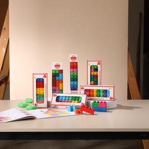 May include: A collection of rectangular boxes containing colorful plastic building blocks, with some blocks arranged outside the packaging. An open instruction booklet with a drawing of a house sits in front of the boxes on a flat surface.
