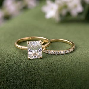 Puede incluir: Un anillo de compromiso de oro con una gran piedra central de corte radiante y una alianza de boda de oro a juego con una hilera de pequeños diamantes de corte redondo. Ambos anillos están hechos de oro amarillo pulido y brillan.