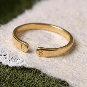 Puede incluir: Un anillo abierto de tono dorado con un acabado liso y pulido. Los dos extremos de la banda están ligeramente aplanados y cada uno está grabado con una letra mayúscula, específicamente la letra E en un lado y la letra S en el otro.
