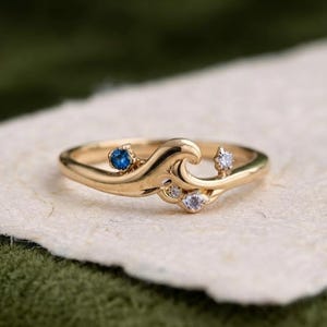 Peut inclure: Une bague en or au design en forme de vague, ornée d'une petite pierre précieuse bleue ronde et de trois pierres précieuses rondes transparentes. L'anneau présente une finition lisse et polie, suivant une silhouette incurvée et fluide.