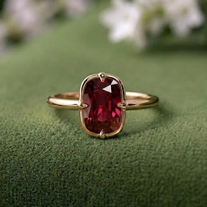 May include: A gold ring featuring a rectangular, cushion-cut deep red gemstone set in a four-prong mounting. The band is smooth and polished, showcasing a simple design that highlights the vibrant colour and faceted surface of the central stone.