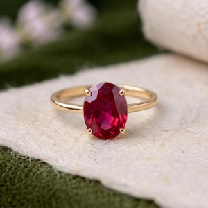 Könnte beinhalten: Ein Goldring mit einem großen, oval geschliffenen, tiefroten Edelstein, der von vier Krappen gehalten wird. Die Ringschiene ist glatt und poliert und zeigt ein schlichtes Design, das die leuchtende Farbe und die facettierte Oberfläche betont.