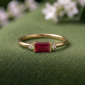 Op de afbeelding: Een gouden ring met een centrale rechthoekige robijnrode edelsteen in baguette-slijpvorm, geflankeerd door twee kleine ronde witte diamanten. De ringband is glad en gepolijst, wat de levendige rode steen en heldere diamanten benadrukt.