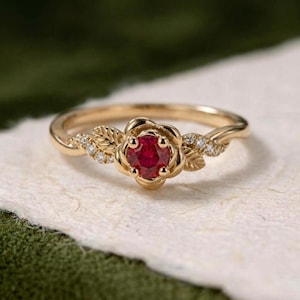 Può includere: Un anello in oro con una pietra preziosa rossa a taglio rotondo incastonata in un design a forma di rosa. La fascia è decorata con piccole pietre chiare e dettagli a forma di foglia scolpita su entrambi i lati dell'elemento centrale.