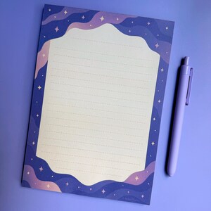 May include: A rectangular notepad with a cream-coloured centre featuring horizontal dashed lines, surrounded by a dark purple border with wavy light purple bands and small four-pointed stars. A light purple retractable ballpoint pen is placed nearby.