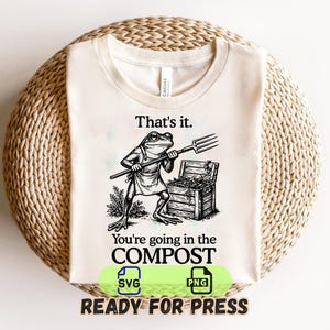 May include: A cream-colored t-shirt featuring a black ink-style illustration of a frog wearing an apron and holding a pitchfork next to a wooden crate. The text reads: That's it. You're going in the COMPOST. The design is centered on the chest.