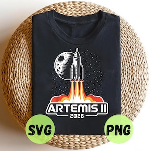May include: Black t-shirt featuring a rocket launching into space with a moon in the background. The design includes the text "ARTEMIS II 2026" in white. The shirt is displayed on a woven surface.