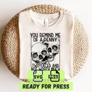 Op de afbeelding: Crèmekleurig T-shirt met een afbeelding van twee schedels en de tekst "YOU REMIND ME OF A PENNY, TWO-FACED AND NOT WORTH MUCH". Inclusief SVG- en PNG-pictogrammen en de woorden "READY FOR PRESS".