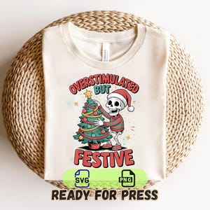 May include: Cream-colored t-shirt with a graphic of a skeleton wearing a Santa hat and sweater, hugging a Christmas tree. The text reads "OVERSTIMULATED BUT FESTIVE". The shirt is on a woven, circular surface.