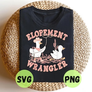 May include: Black t-shirt featuring a cartoon duck dressed as a cowboy with the text "ELOPEMENT WRANGLER" in pink. The design includes a cactus and a second duck. The shirt is on a woven surface.