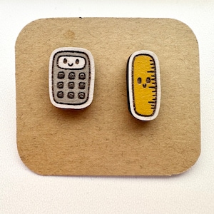 Peut inclure: Une paire de boucles d'oreilles en bois avec un design de calculatrice et de règle. La calculatrice est grise avec un visage blanc et un visage souriant. La règle est jaune avec un visage blanc et un visage souriant.