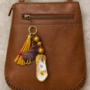 May include: Brown leather crossbody bag with a gold zipper and a decorative charm. The charm features a yellow tassel, a rooster pendant with red and black accents, and a painted oyster shell.