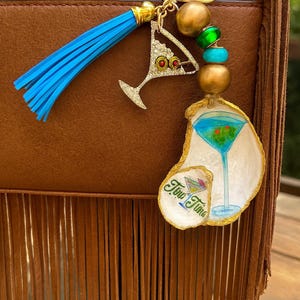 May include: A gold keyring with a blue tassel, gold beads, and a martini charm. The keyring also features a painted oyster shell with a martini design and the text "Two Time".