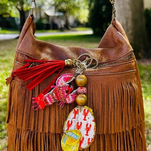 Pode incluir: Bolsa de mão de couro marrom com franjas e alça de contas. A bolsa apresenta uma borla vermelha e um pingente de lagosta com detalhes dourados. Um pequeno pingente com o texto "ORME" também está anexado.
