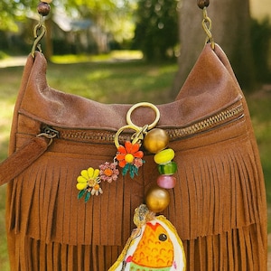 May include: Brown leather fringe bag with a beaded strap and a decorative keychain. The keychain features colorful flower charms, gold beads, and a painted oyster shell. The bag has a zipper closure and a long shoulder strap.
