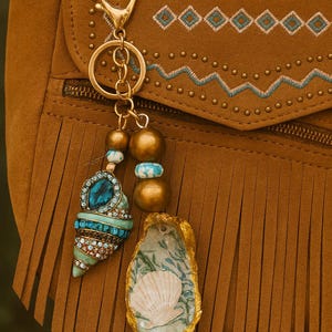May include: A gold-coloured keyring with a seashell charm and a decorative pendant. The pendant features a turquoise and crystal-studded seashell design, along with gold beads and a painted oyster shell. The background is a brown suede bag with fringe.