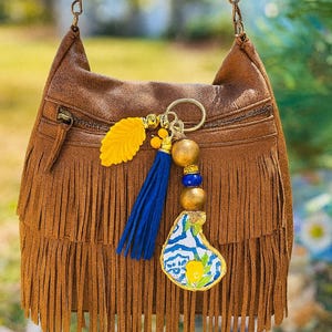 May include: Brown suede fringe bag with a beaded strap. The bag features a zipper closure and a decorative keychain with a yellow leaf, blue tassel, wooden beads, and a patterned charm. The bag is designed with a bohemian style.
