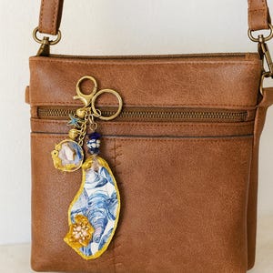 May include: A brown leather crossbody bag with a decorative keychain. The keychain features a blue and white patterned oyster shell charm, a small gold-framed pendant, and blue bead accents. The bag has a zippered pocket.