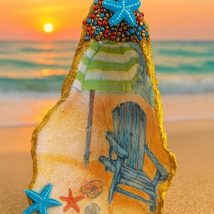 May include: A decorative seashell with a beach scene. The shell features a painted beach chair, umbrella, and starfish. The top is adorned with blue and red beads and a blue starfish. The edges are trimmed in gold.