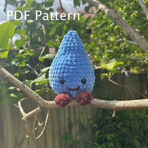 May include: A blue crocheted raindrop toy with a smiling face and small brown feet, sitting on a tree branch. The text "PDF Pattern" is visible. The background features green leaves and branches, creating a natural setting.