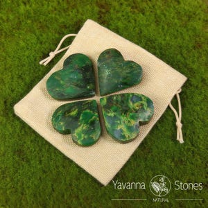 May include: Four heart-shaped green stones with a marbled pattern, arranged on a beige drawstring bag. The stones are a deep green with lighter green and yellow veining. The bag sits on a bed of green moss. The text "Yavanna Stones NATURAL" is visible.