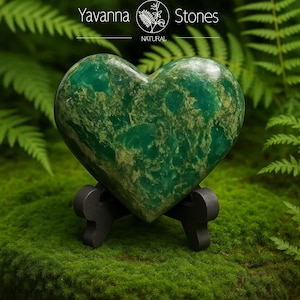 May include: A heart-shaped green stone with gold veining, displayed on a small black stand. The stone is set against a backdrop of green moss and ferns. The text "Yavanna Stones NATURAL" is visible in the upper portion of the image.