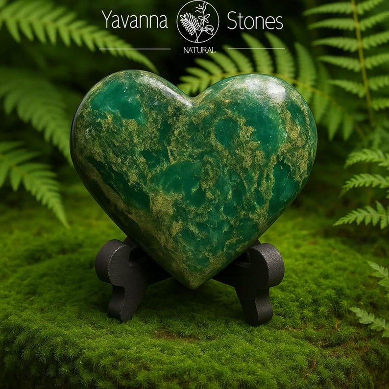 May include: A heart-shaped green stone with gold veining, displayed on a small black stand. The stone is set against a backdrop of green moss and ferns. The text "Yavanna Stones NATURAL" is visible in the upper portion of the image.