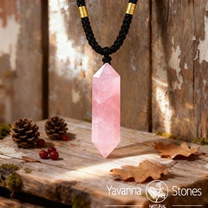 May include: A pink rose quartz crystal pendant necklace with a black braided cord and gold accents. The crystal is a long, hexagonal shape. The necklace is displayed on a wooden surface with pinecones, berries, and leaves. The text "Yavanna Stones" is visible.