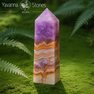 May include: A rectangular amethyst crystal obelisk with a pointed top. The stone features layers of purple, orange, and white hues. The obelisk is set against a green, mossy background. The text "Yavanna Stones NATURAL" is at the top.
